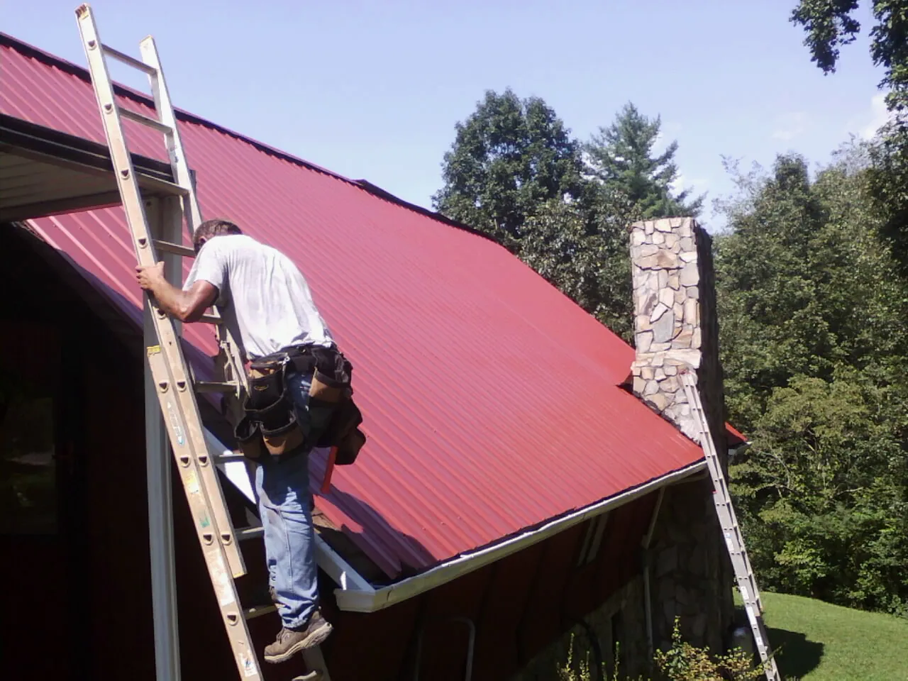 Roofer on a ladder beside a red metal roof and stone chimney — Soffit & Fascia Repair by Roofing Brothers in Annapolis Junction